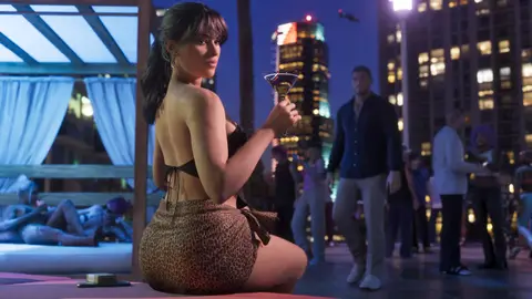 A woman with a cocktail at a rooftop pool party above the night skyline