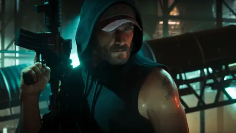 Jason in a hood and cap holding a carbine upright in a dark industrial yard