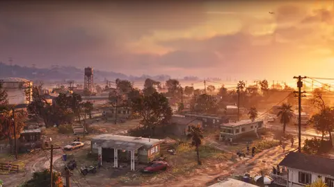 A trailer town of shacks and pickups under power lines at sunset, storage tanks and a water tower behind